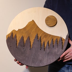 May include: A circular wooden wall art piece featuring a layered mountain landscape. The design includes a light wood background, a stained brown mountain peak, a dark brown forest silhouette in the foreground, and a raised circular brown sun.