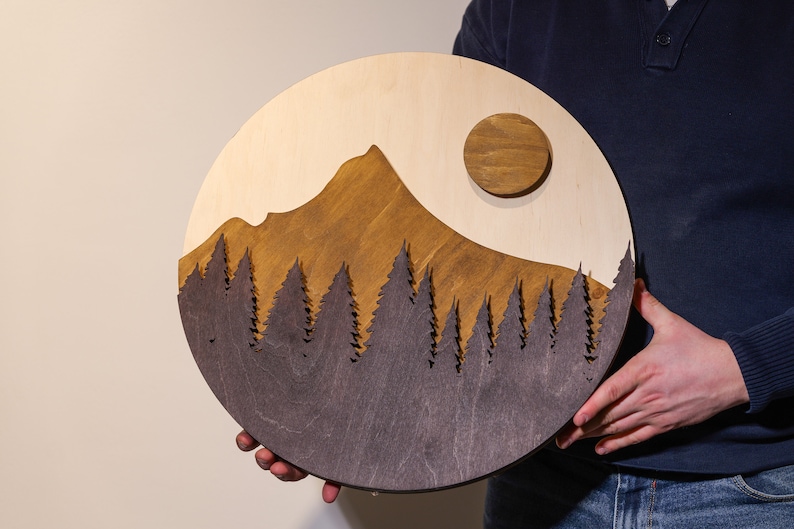 May include: A circular wooden wall art piece featuring a layered mountain landscape. The design includes a light wood background, a stained brown mountain peak, a dark brown forest silhouette in the foreground, and a raised circular brown sun.