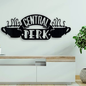 以下が含まれることがあります： テレビ番組Friendsの「Central Perk」ロゴをあしらった黒い金属製のウォールアート。デザインには、湯気の立つコーヒーカップ2つと、「CENTRAL PERK」の文字がバナーで描かれています。リビングルームやコーヒーショップの装飾品です。