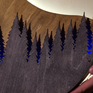 May include: A circular wooden wall art piece featuring a laser-cut silhouette of a dense pine forest in dark brown wood. The cutout design reveals a vibrant blue light emanating from behind the trees, creating a high-contrast visual effect.