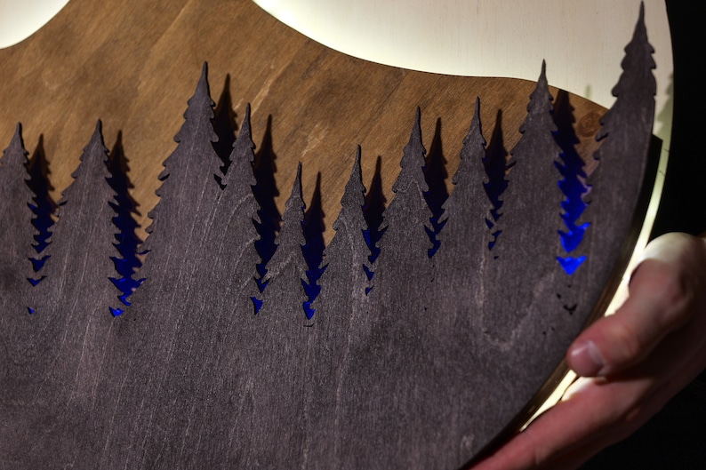 May include: A circular wooden wall art piece featuring a laser-cut silhouette of a dense pine forest in dark brown wood. The cutout design reveals a vibrant blue light emanating from behind the trees, creating a high-contrast visual effect.