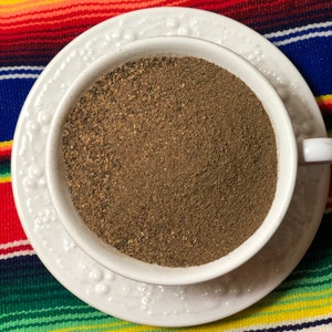 Puede incluir: Una taza de té de cerámica blanca llena de un polvo marrón, probablemente café molido o especias, sobre un platillo blanco con un diseño floral. El platillo está sobre un fondo de tela de rayas de colores.