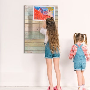 May include: Two children, one with long brown hair and one with short brown hair, are hanging a framed artwork on a wall. The artwork is a colorful painting on a wooden background. The artwork is framed with a clear plastic frame.
