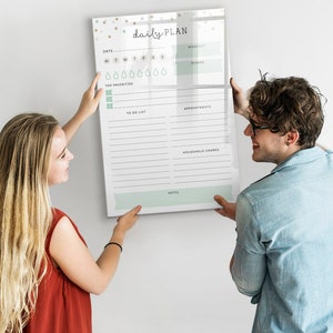 Op de afbeelding: Een stel hangt een witte dagelijkse planner op een witte muur. De planner heeft een lichtblauw en groen ontwerp met de woorden "daily PLAN" bovenaan. De planner heeft secties voor de dagen van de week, topprioriteiten, to-do-lijst, afspraken, huishoudelijke taken en notities.