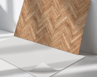 May include: A rectangular sheet featuring a light brown wood grain pattern arranged in a herringbone design. The sheet has a smooth, white adhesive backing that is partially peeled back at one corner, revealing the underside of the material.