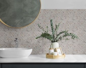 May include: A white oval vessel sink with a chrome faucet, a round mirror with a gold frame, and a light-colored terrazzo wall. A white ceramic soap dispenser and a small wooden picture frame sit on a round woven tray with green eucalyptus.