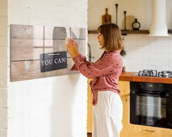 May include: A woman hangs a wooden sign with the text "YOU CAN!" on a white brick wall. The sign is hanging from a string and has a rustic woodgrain background.