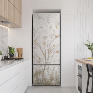 Può includere: Un frigorifero con un design floreale personalizzato. Il design presenta un albero con fiori bianchi e foglie color oro su uno sfondo grigio chiaro. Il frigorifero si trova in un ambiente cucina moderno con mobili e piani di lavoro bianchi.