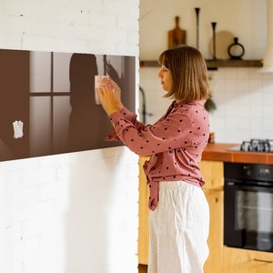 Op de afbeelding: Een vrouw in een roze gebloemd shirt en een witte broek maakt een bruin glazen magnetisch whiteboard schoon met een schoonmaakdoek.