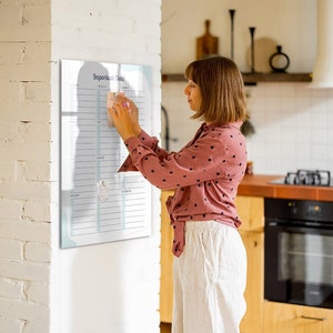 May include: A woman wearing a pink polka dot shirt and white trousers is adding a sticky note to a white dry erase board with the words "Important Tasks" at the top. The board is hanging on a white brick wall.