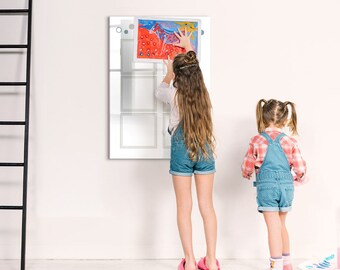 May include: Two children, one wearing a blue and white plaid shirt and denim overalls and the other wearing a white shirt and denim shorts, are standing in front of a white wall with a framed picture of a colorful abstract painting.