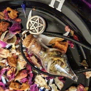 May include: A silver pentagram pendant necklace with a clear glass bottle filled with dried herbs and a cork stopper. The necklace is on a black cord and is lying on a surface with dried rose petals.