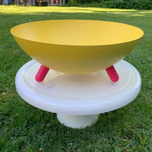 Puede incluir: Un cuenco amarillo con tres patas rosas, apoyado sobre un pedestal blanco. El cuenco tiene un diseño ancho y poco profundo. El pedestal es redondo y blanco. El conjunto está sobre una superficie de hierba verde.