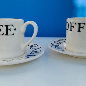 May include: Two off-white ceramic coffee cups with matching saucers. Each cup features the black text "COFFEE" on the side. The saucers are decorated with the blue text "COSTA" and "ARABICA" printed in a circular pattern around the rim.