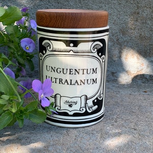 Puede incluir: Tarro cilíndrico de cerámica blanca con tapa de madera. Presenta elementos decorativos negros y el texto "UNGENTUM ULTRALANUM" y "Schering". Flores moradas y hojas verdes en primer plano.