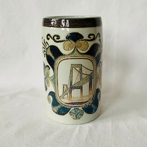 Könnte beinhalten: Eine weiße Keramiktasse mit braunem Rand und einem blauen und braunen Design mit einer Brücke. Das Design ist von einem dekorativen Rand umgeben.