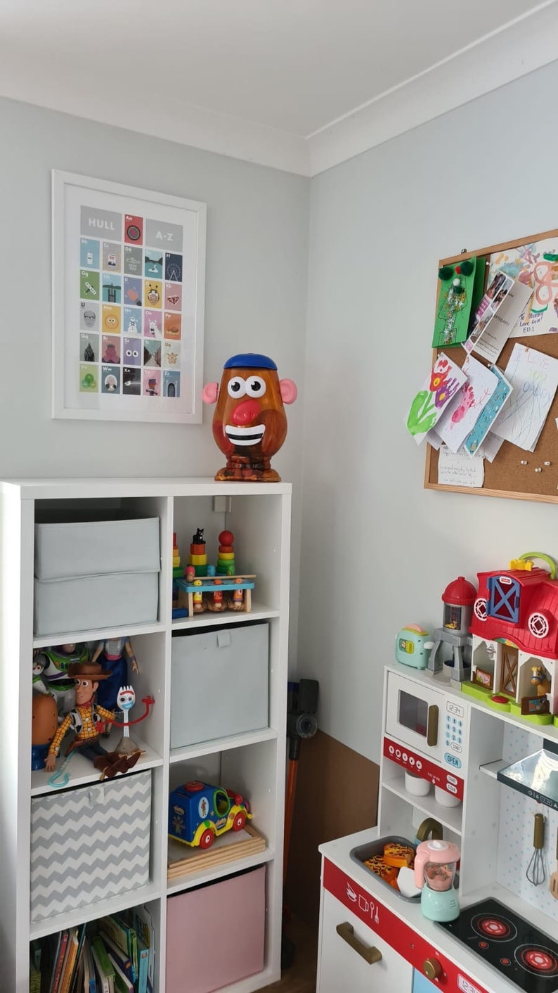 May include: A white bookshelf with toys and books. The top shelf has a Mr. Potato Head toy. The second shelf has a wooden toy stacking tower. The third shelf has a grey storage bin. The fourth shelf has a grey storage bin. The fifth shelf has a toy car. The sixth shelf has a pink storage bin. The bottom shelf has books.