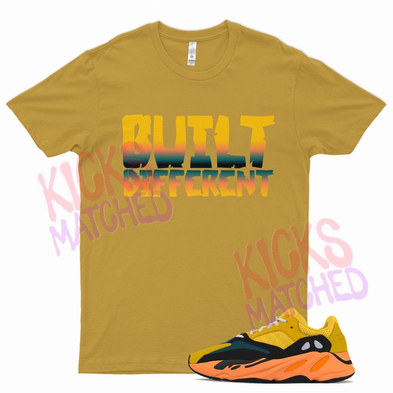 Puede incluir: Una camiseta amarilla mostaza con el texto "BUILT DIFFERENT" en un degradado de amarillo, naranja y azul. La camiseta se combina con un par de zapatillas amarillas, naranjas y negras.