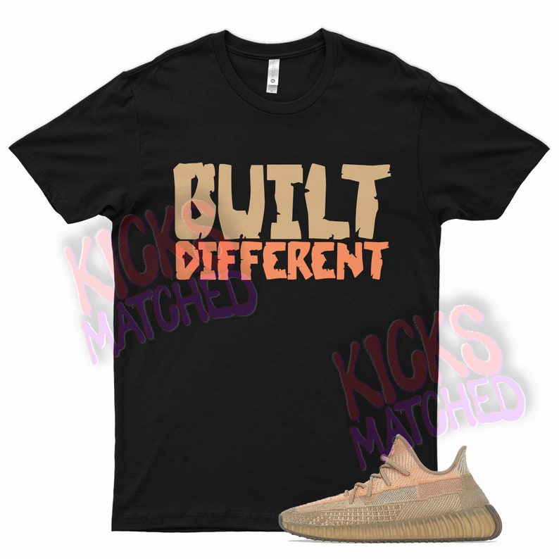 May include: Black t-shirt with the text "BUILT DIFFERENT" in brown and orange lettering. A pair of tan and brown trainers are shown below the shirt.