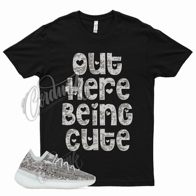 May include: Black t-shirt with the phrase "OUT HERE BEING CUTE" in white, pixelated font. The design includes heart shapes. A pair of gray and white sneakers with a pixelated pattern is in the foreground.