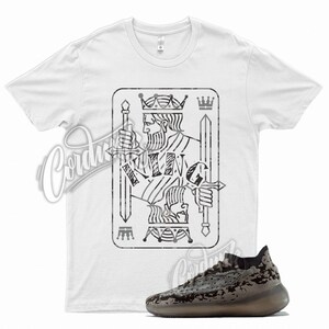 May include: A white t-shirt with a black and white graphic of a king of spades playing card. The graphic has a distressed look. A pair of black and grey sneakers with a camouflage pattern are also pictured.