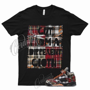 May include: Black t-shirt with a plaid design and the text "CUT FROM A DIFFERENT CLOTH". A pair of plaid sneakers with orange laces are in the lower right corner. The design features scissors and a mix of red, green, and orange plaid patterns.