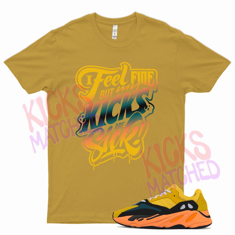 Op de afbeelding: Een mosterdgeel T-shirt met de tekst "I Feel Fine But My Kicks Are Sick!" gedrukt op de voorkant. Een paar gele, zwarte en oranje sneakers zijn te zien onder het T-shirt.