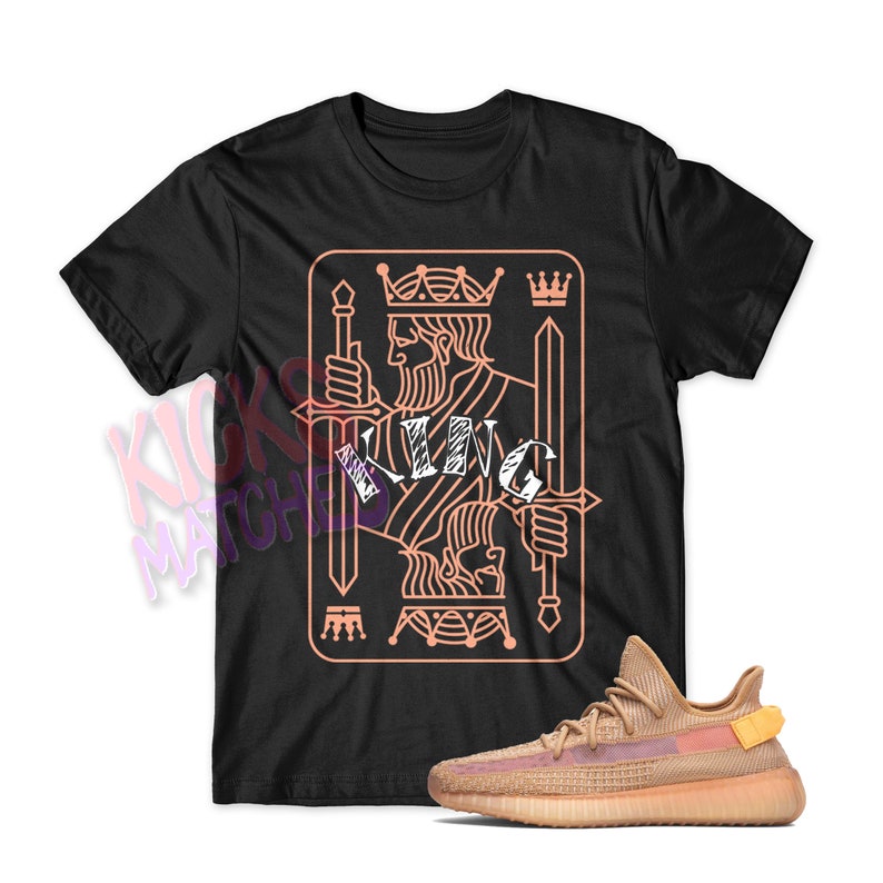 May include: Black t-shirt with a peach-colored king of spades playing card graphic. The word "KING" is written in white across the card. A pair of peach-colored sneakers are in the bottom right corner of the image.