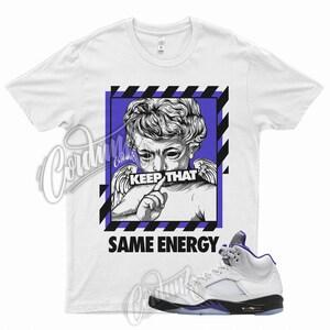 May include: White t-shirt with a graphic of an angel figure with the text "KEEP THAT" and "SAME ENERGY". A white and purple sneaker is next to the shirt.