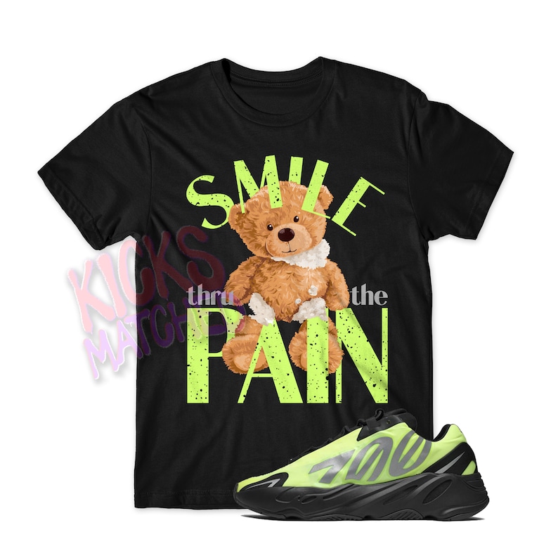 以下が含まれることがあります： 黒いTシャツに、包帯をしたテディベアと「Smile thru the pain」というテキストがネオングリーンで描かれたグラフィックがプリントされています。黒とネオングリーンのスニーカーも写っています。