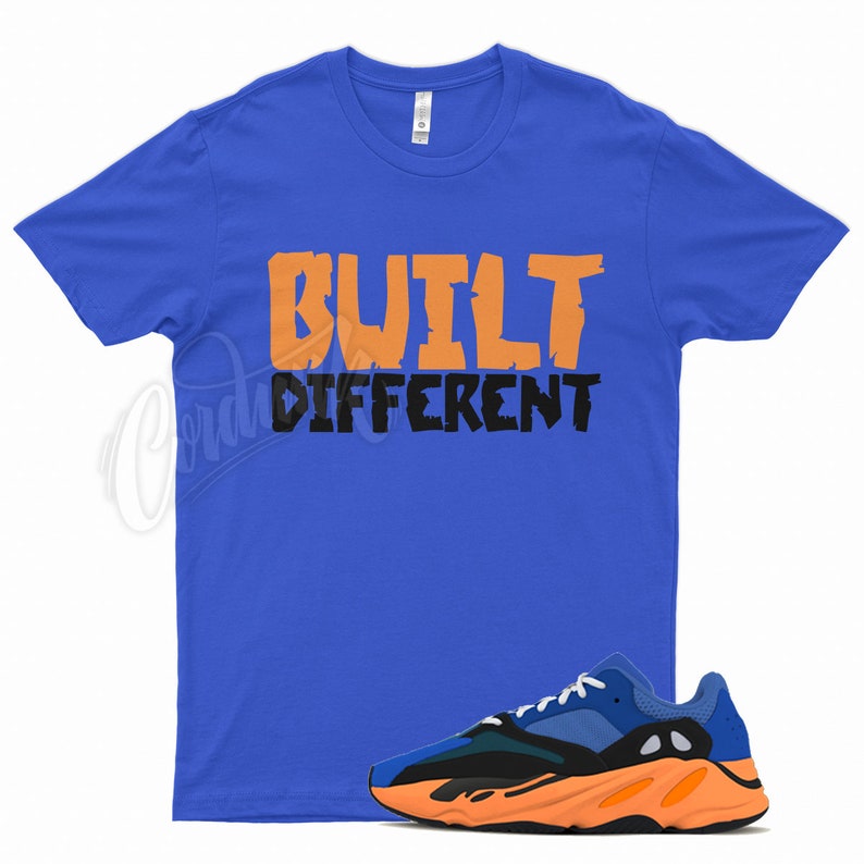Puede incluir: Una camiseta azul real con las palabras "BUILT DIFFERENT" en naranja y negro. Un par de zapatillas deportivas azules, negras y naranjas se encuentra en la parte inferior de la imagen.