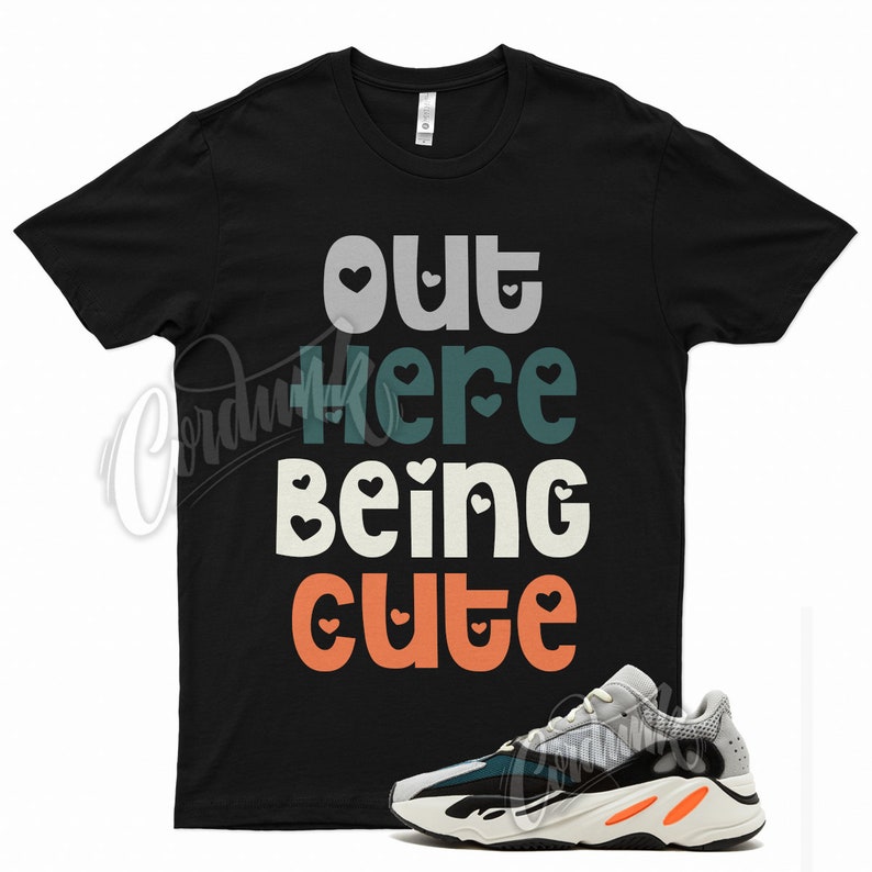 May include: Black t-shirt with the text "out here being cute" in colorful letters with small hearts around each letter. A pair of white, black, and green sneakers with orange accents are shown below the shirt.