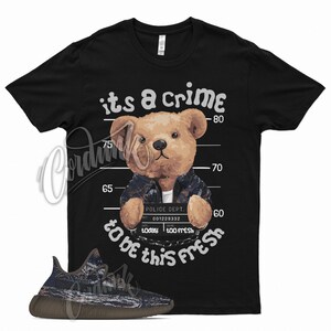 May include: Black t-shirt with a teddy bear mugshot design. The bear wears a jacket and holds a sign that reads "POLICE DEPT." and "too fresh." The text "its a crime to be this fresh" is above and below the bear. A sneaker is in the foreground.