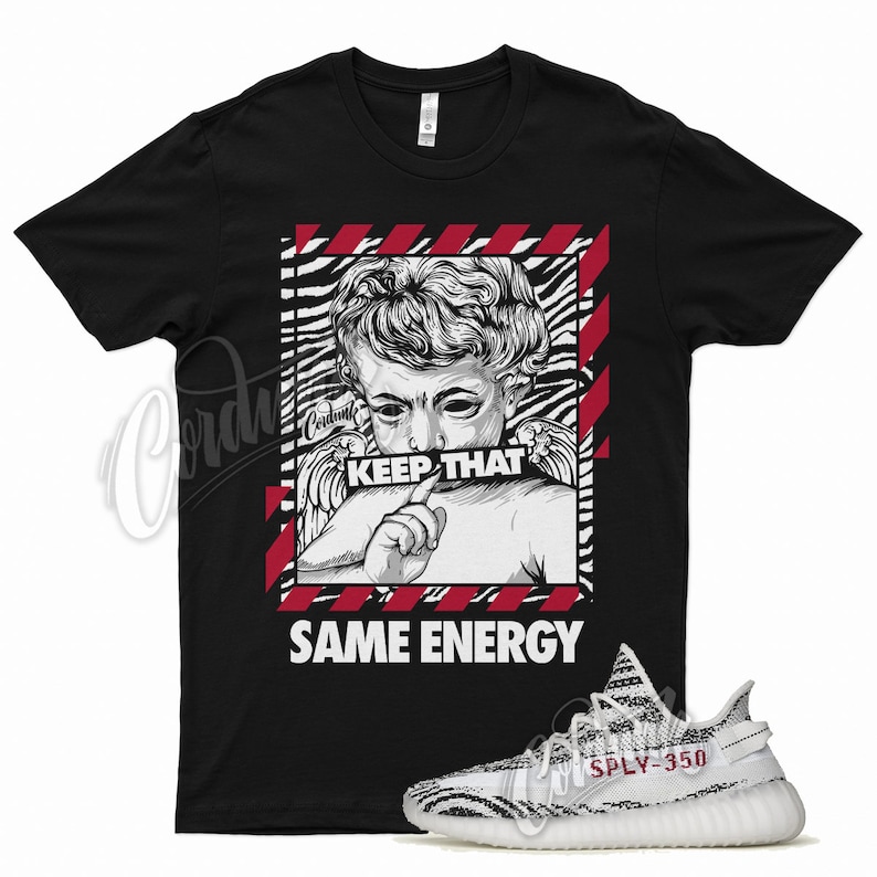 Pu&ograve; includere: T-shirt nera con grafica di statua e la scritta "KEEP THAT SAME ENERGY". Un paio di scarpe da ginnastica bianche e nere con la scritta "SPY-350" sono nell'immagine.