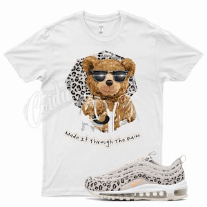 May include: White t-shirt with a cartoon bear wearing sunglasses and a leopard print jacket. The text "Made it through the rain" is printed below the bear. A pair of white and beige Nike Air Max 97 sneakers with a leopard print design are shown below the t-shirt.