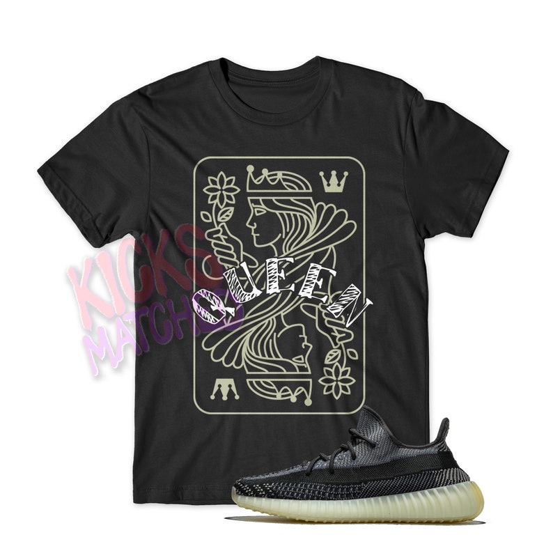 May include: Black t-shirt with a graphic of a queen of spades playing card. The word "Queen" is written over the graphic. A pair of black and white sneakers are in the foreground.