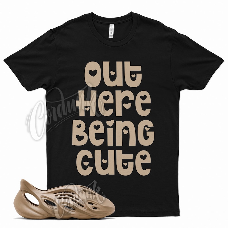 May include: A black t-shirt with the text "out here being cute" in brown with small hearts around the letters. A pair of brown foam clogs are in the bottom of the image.