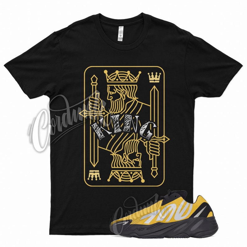 May include: Black t-shirt with a gold king of spades playing card graphic. The shirt is paired with black and yellow sneakers.