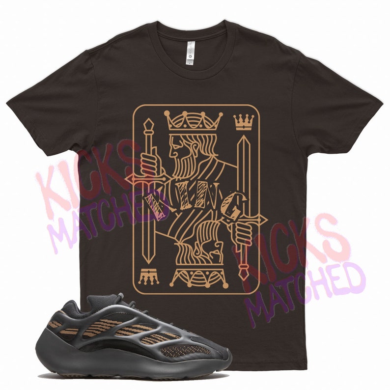 May include: A brown t-shirt with a gold outline of a king of spades playing card. The king is holding two swords. The t-shirt is paired with black and brown sneakers.