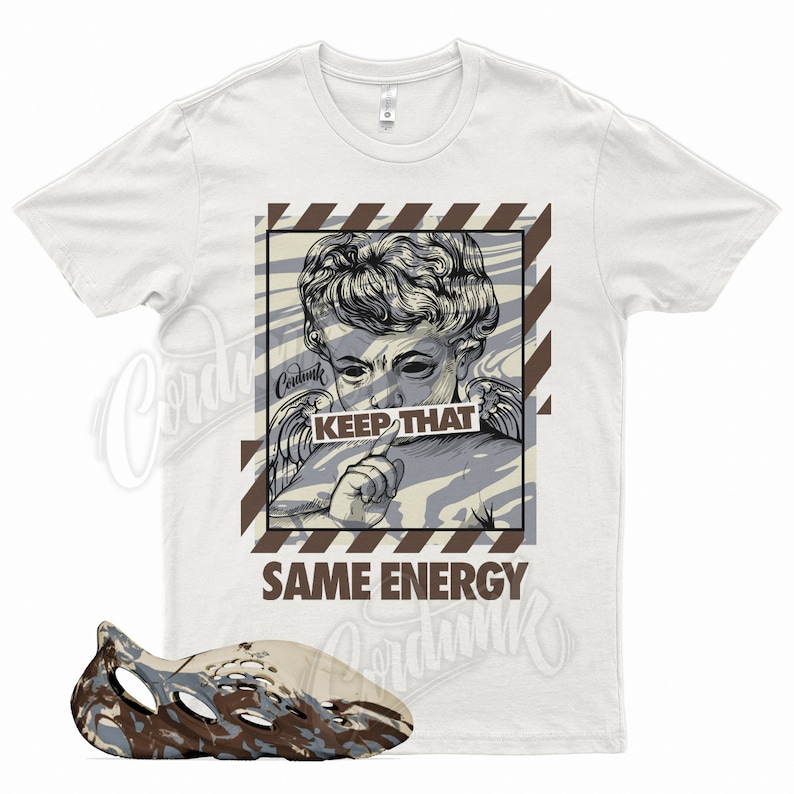 Pu&ograve; includere: T-shirt bianca con grafica di angelo e la scritta "KEEP THAT" e "SAME ENERGY". Un paio di scarpe da ginnastica in schiuma marrone, beige e blu in primo piano.