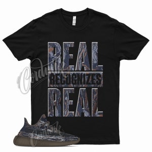 May include: Black t-shirt with the text "REAL recognizes REAL" in a camouflage pattern. The shirt is paired with a pair of brown and blue sneakers with a camouflage pattern.