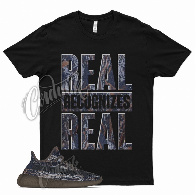 K&ouml;nnte beinhalten: Schwarzes T-Shirt mit dem Text "REAL recognizes REAL" in einem Tarnmuster. Das Shirt wird mit einem Paar braunen und blauen Sneakern mit Tarnmuster kombiniert.