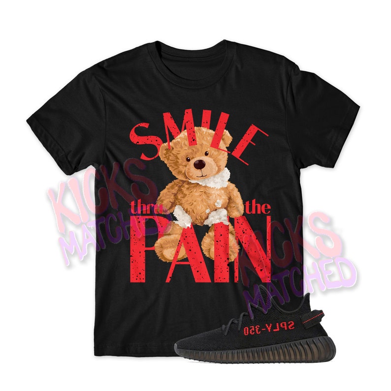 Pu&ograve; includere: Maglietta nera con una grafica rossa di un orsacchiotto di peluche con un bendaggio sul braccio. La grafica dice "Smile thru the pain". Sono anche raffigurate un paio di scarpe Yeezy nere.
