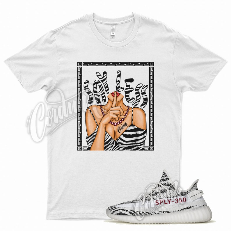 以下が含まれることがあります： ゼブラ柄のドレスを着た女性のグラフィックが描かれた白いTシャツで、その上に「SAY LESS」という言葉が書かれています。 「SPLY-350」の文字が入った白と黒のスニーカーが前景にあります。