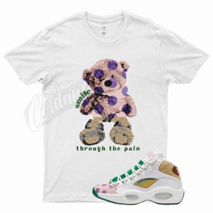 May include: White t-shirt with a cartoon teddy bear wearing purple polka dots and the text "smile through the pain". A pair of white, green, and pink sneakers with a brown and white patterned design.