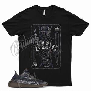 May include: Black t-shirt with a graphic of a king of spades playing card with the word "KING" written on it. A pair of brown and grey sneakers are in the foreground.