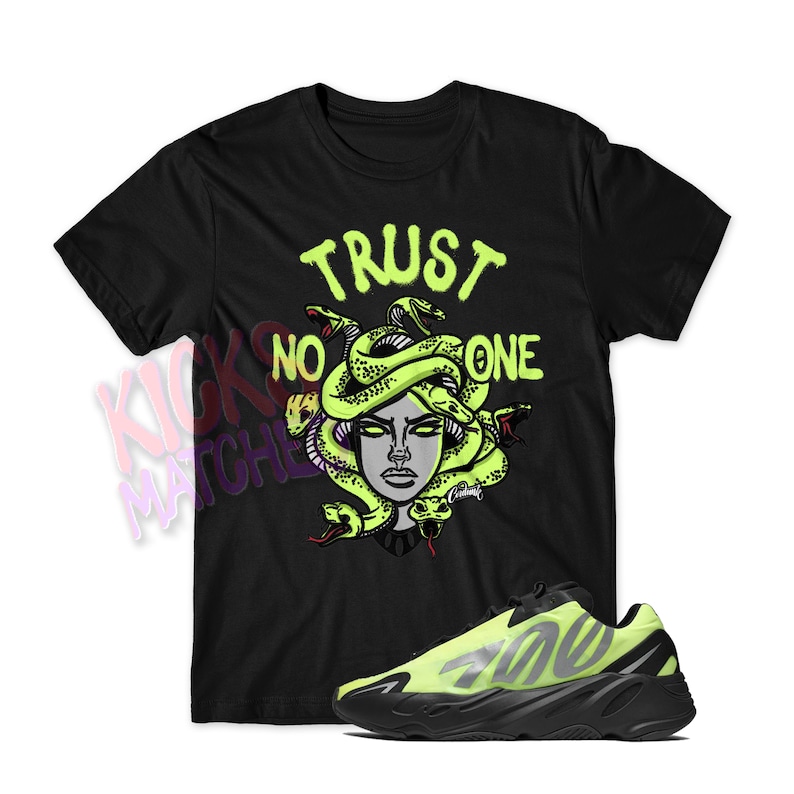 以下が含まれることがあります： 黒いTシャツに、ヘビが絡み付いたメデューサの頭と「TRUST NO ONE」の文字が緑色のグラフィックで描かれている。Tシャツの下には、黒と蛍光緑のスニーカーが置かれている。