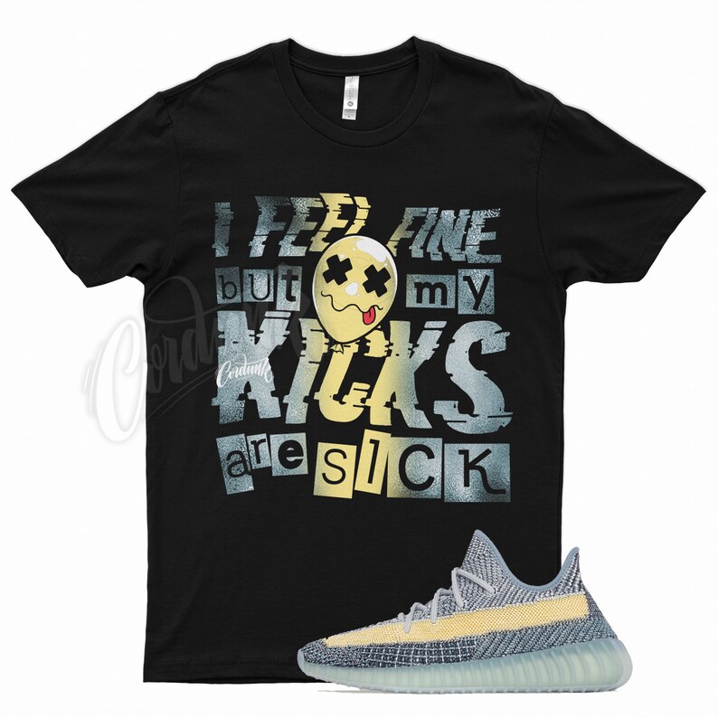 May include: Black t-shirt with a graphic print that says "I feel fine but my kicks are sick". The graphic features a yellow smiley face with X's for eyes and a tongue sticking out. A pair of light blue and yellow sneakers are shown below the t-shirt.