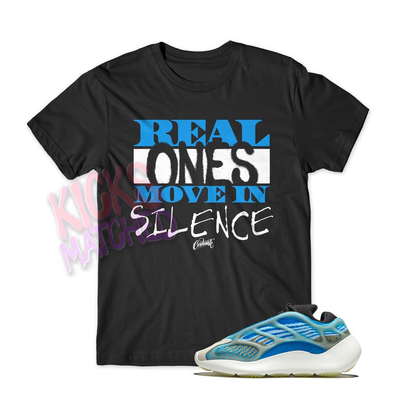 May include: A black t-shirt with the text "REAL ONES MOVE IN SILENCE" in blue and white lettering. The shirt is paired with a pair of white and blue trainers.