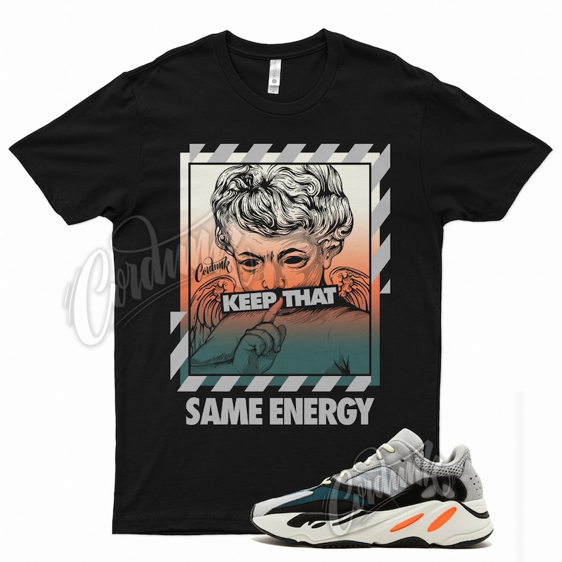 May include: Black t-shirt featuring a graphic of a statue with the text "KEEP THAT SAME ENERGY." A pair of gray, black, and orange sneakers are in the foreground. The shirt has a crew neck.